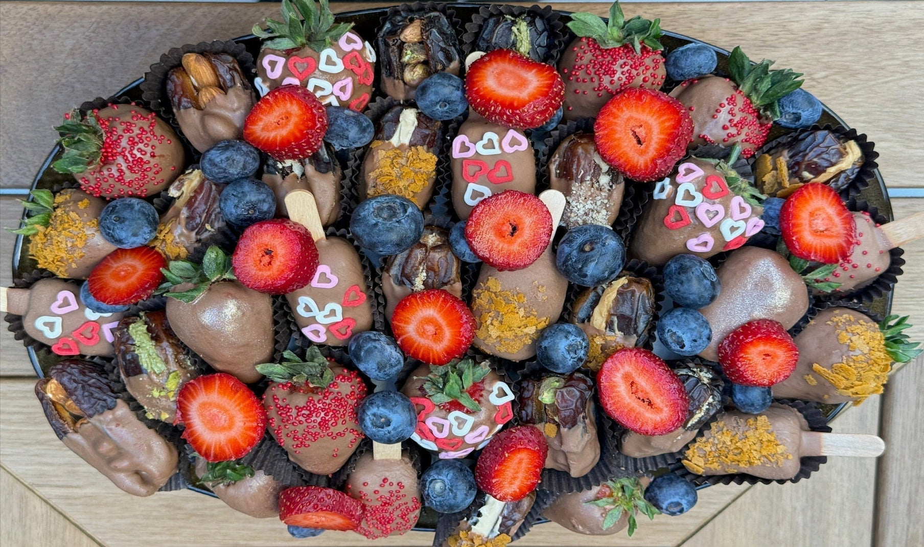 A top-down view of a large, oval-shaped chocolate and fruit snack tray filled with chocolate-covered strawberries, blueberries, dates, and chocolate popsicles decorated with hearts and sprinkles, set on a light wood background.