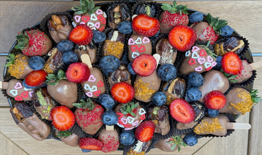 A top-down view of a large, oval-shaped chocolate and fruit snack tray filled with chocolate-covered strawberries, blueberries, dates, and chocolate popsicles decorated with hearts and sprinkles, set on a light wood background.